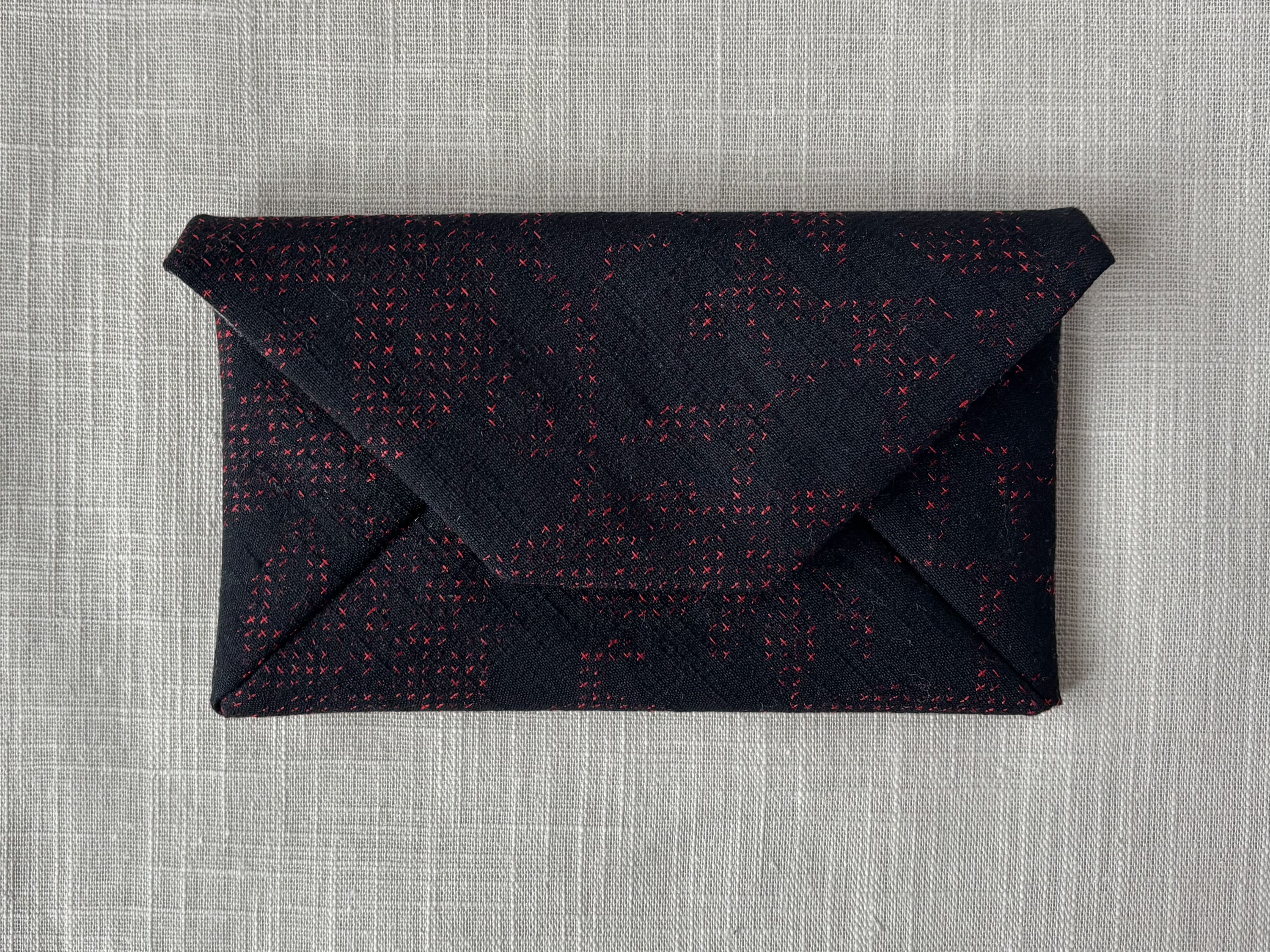 Front view of the matte black sukiya bag with crimson cross stitch pattern on a cream coloured textured linen background.